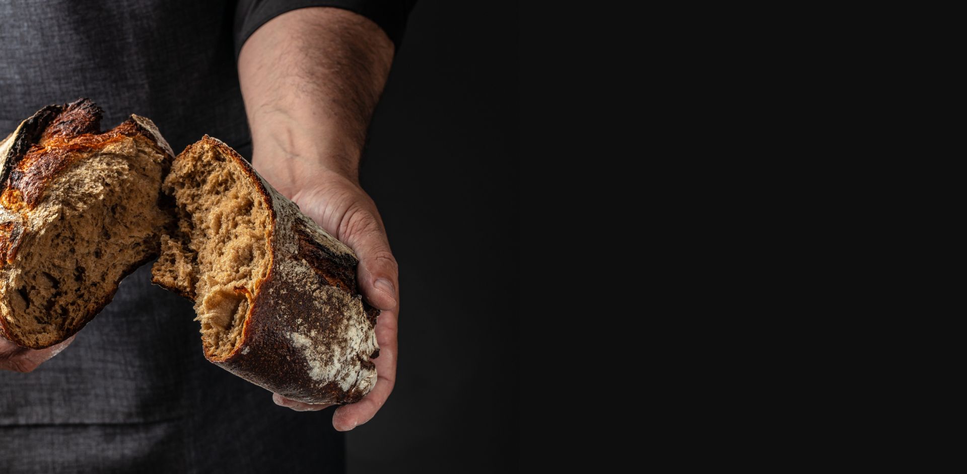 Zwei Hände halten ein aufgebrochenes, rustikales Brot vor dunklem Hintergrund